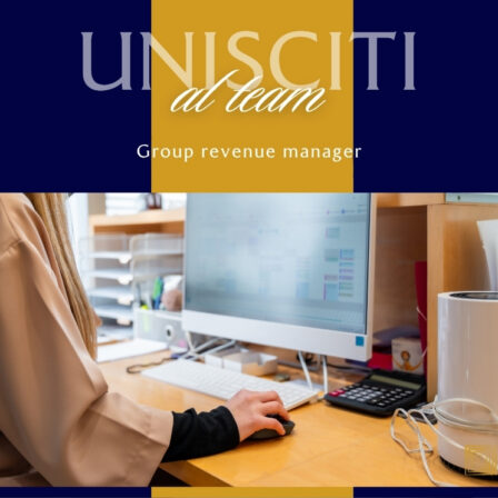 Una persona seduta a una scrivania d'ufficio che lavora su un grande computer desktop con tastiera; sulla scrivania si vedono una calcolatrice, una tazza e cavi, in un ambiente con pareti blu scuro e una fascia gialla al centro.